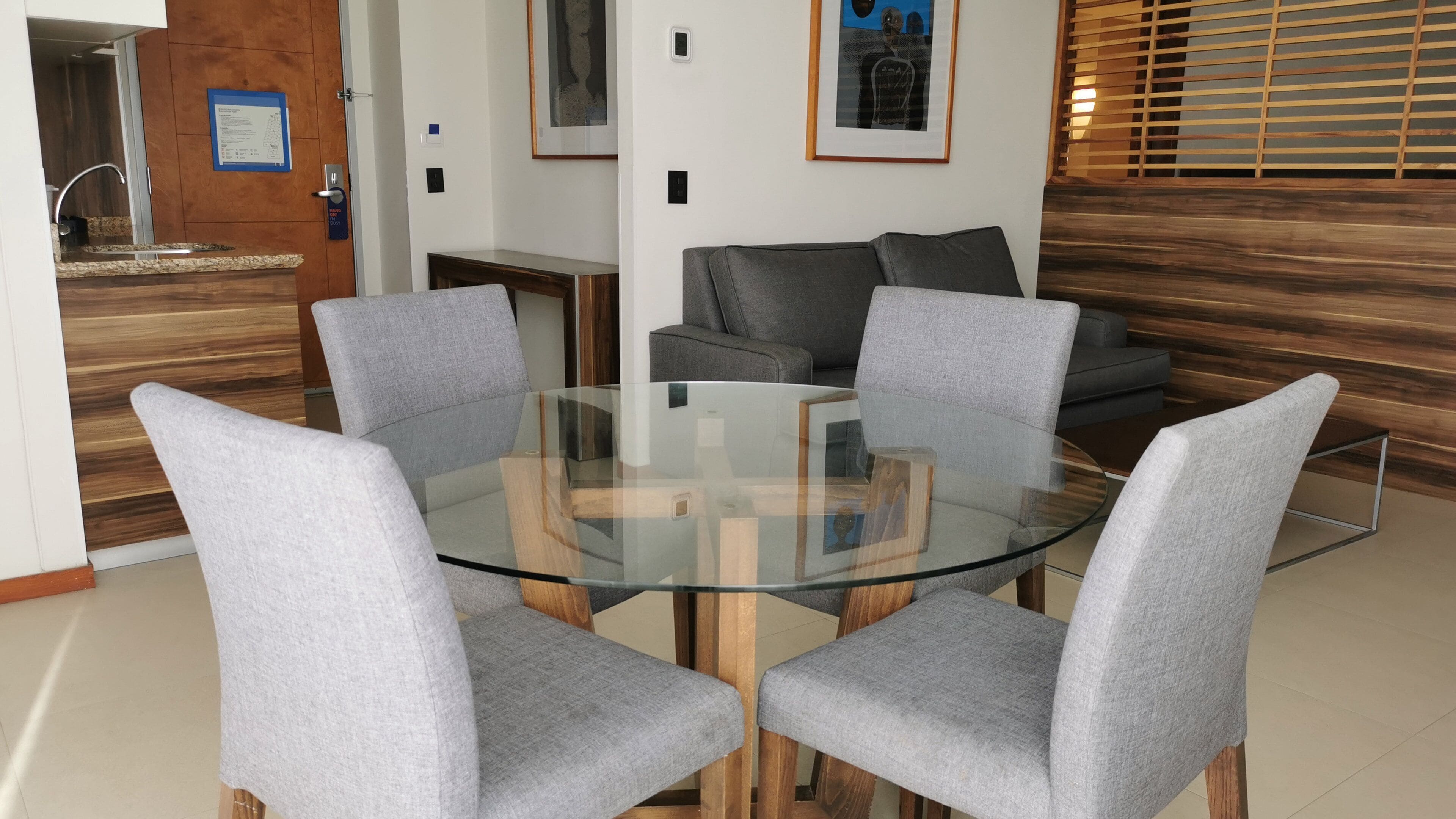 Un salon moderne avec une table basse au plateau en verre, entourée de quatre fauteuils gris à revêtement. Un canapé gris est placé contre le mur, et un panneau décoratif en bois est visible.