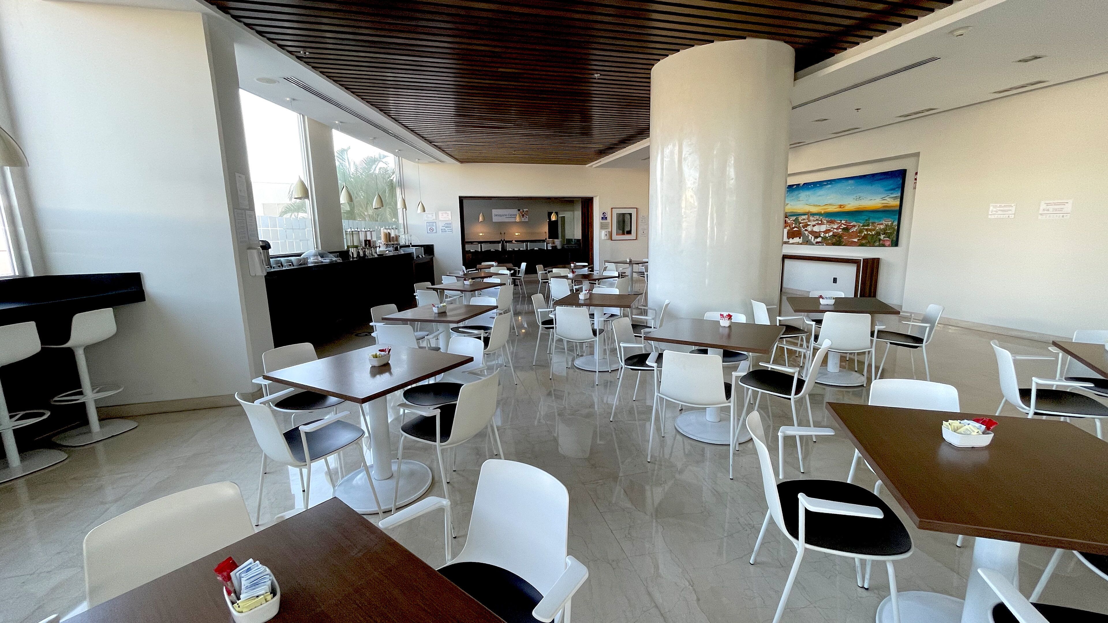 A spacious dining area with multiple tables and chairs, a counter with a menu, and a view of the cityscape through large windows.