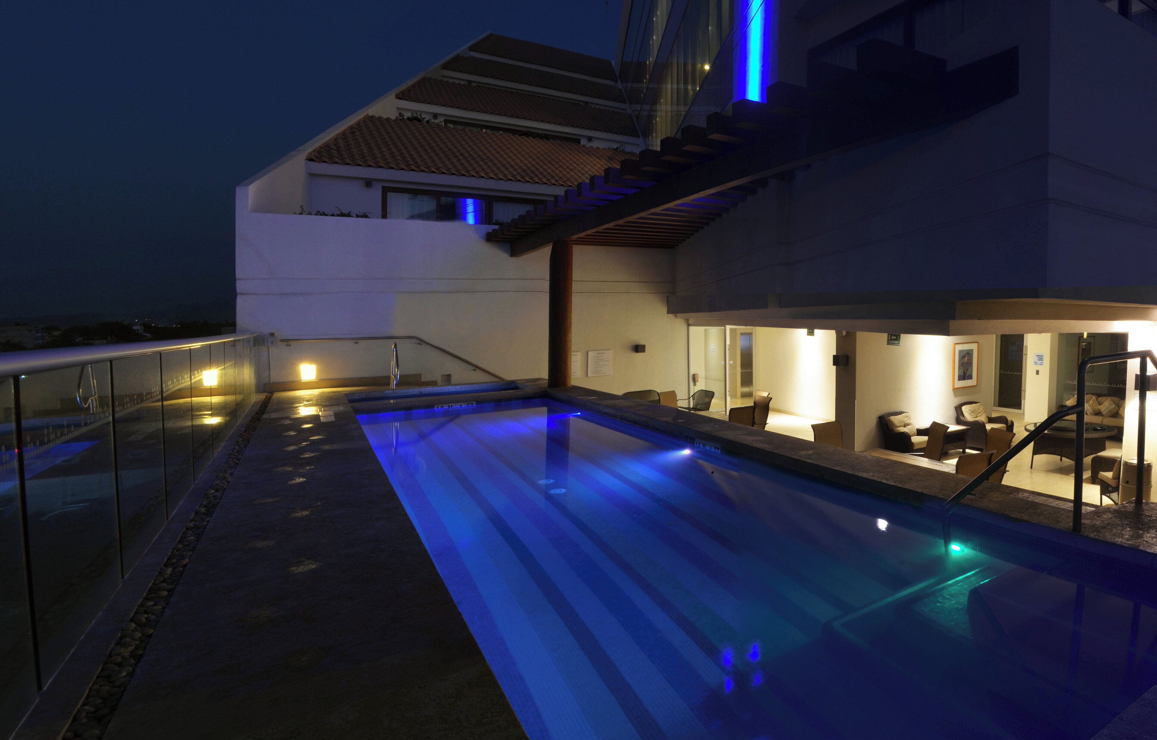 Una piscina moderna al aire libre con barandilla de cristal, una escalera que conduce a un nivel superior y un área de descanso con sillas y una mesa.