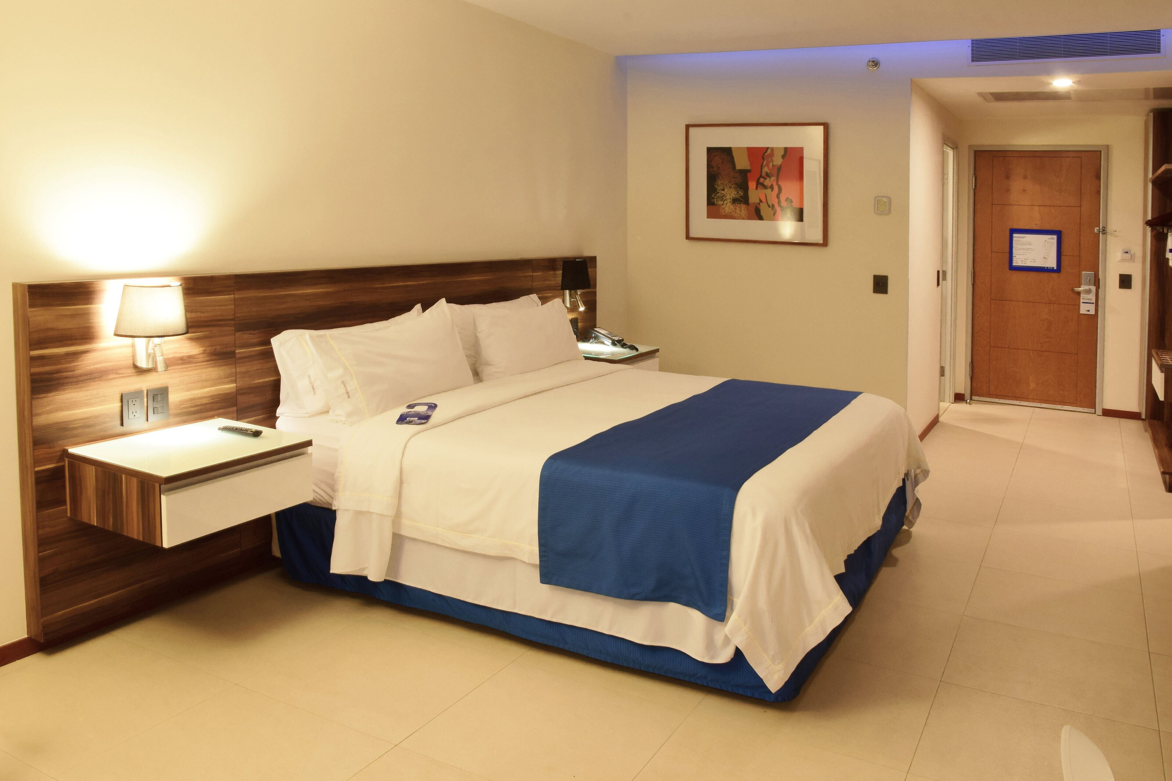 A hotel room with a large bed, bedside tables, a lamp, a painting on the wall, and a door.
