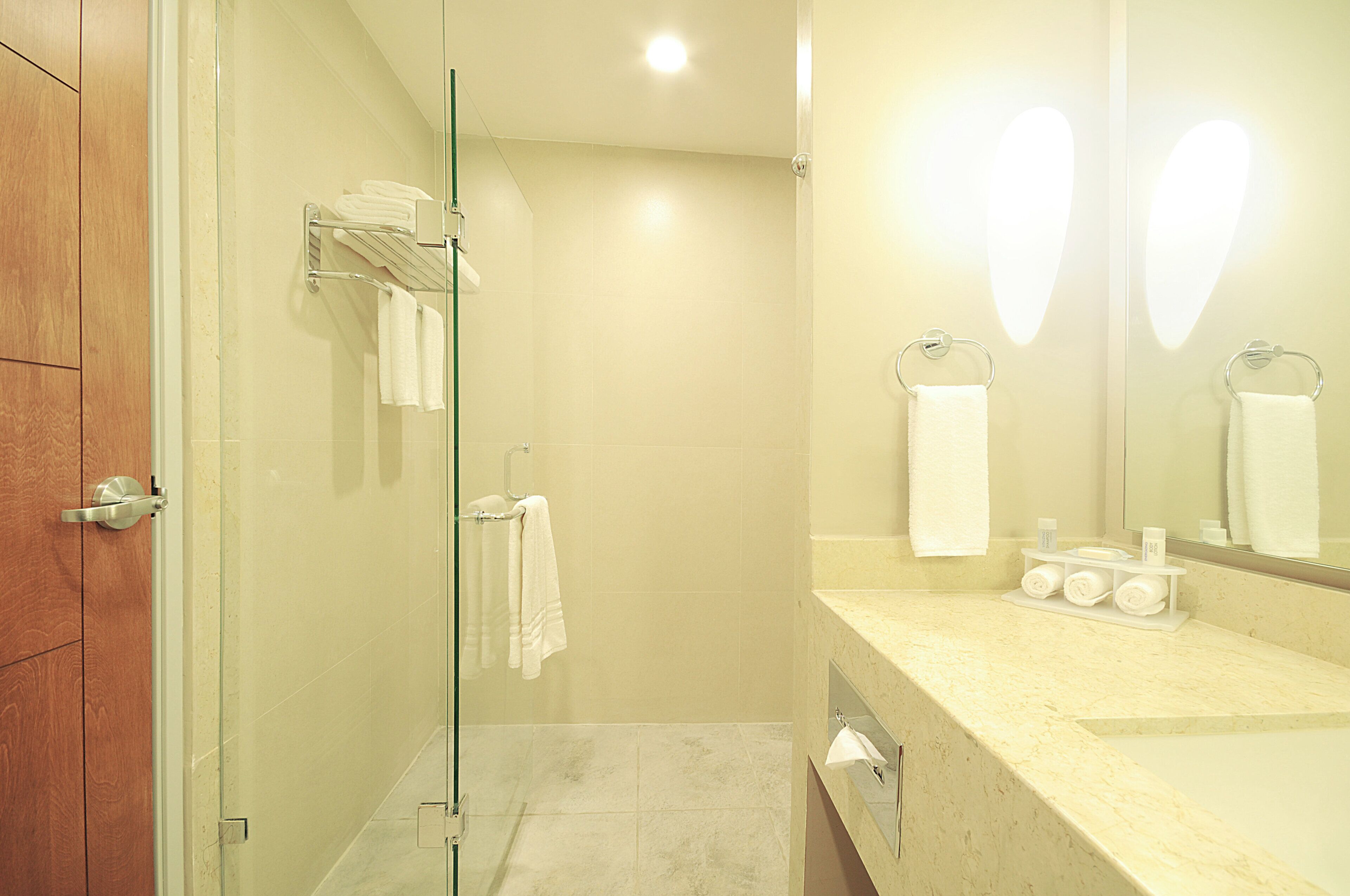 Un bany d'hotel amb un envà d'aixeta de vidre, un lavabo amb productes de tocador i un mirall amb llums de forma de cor.