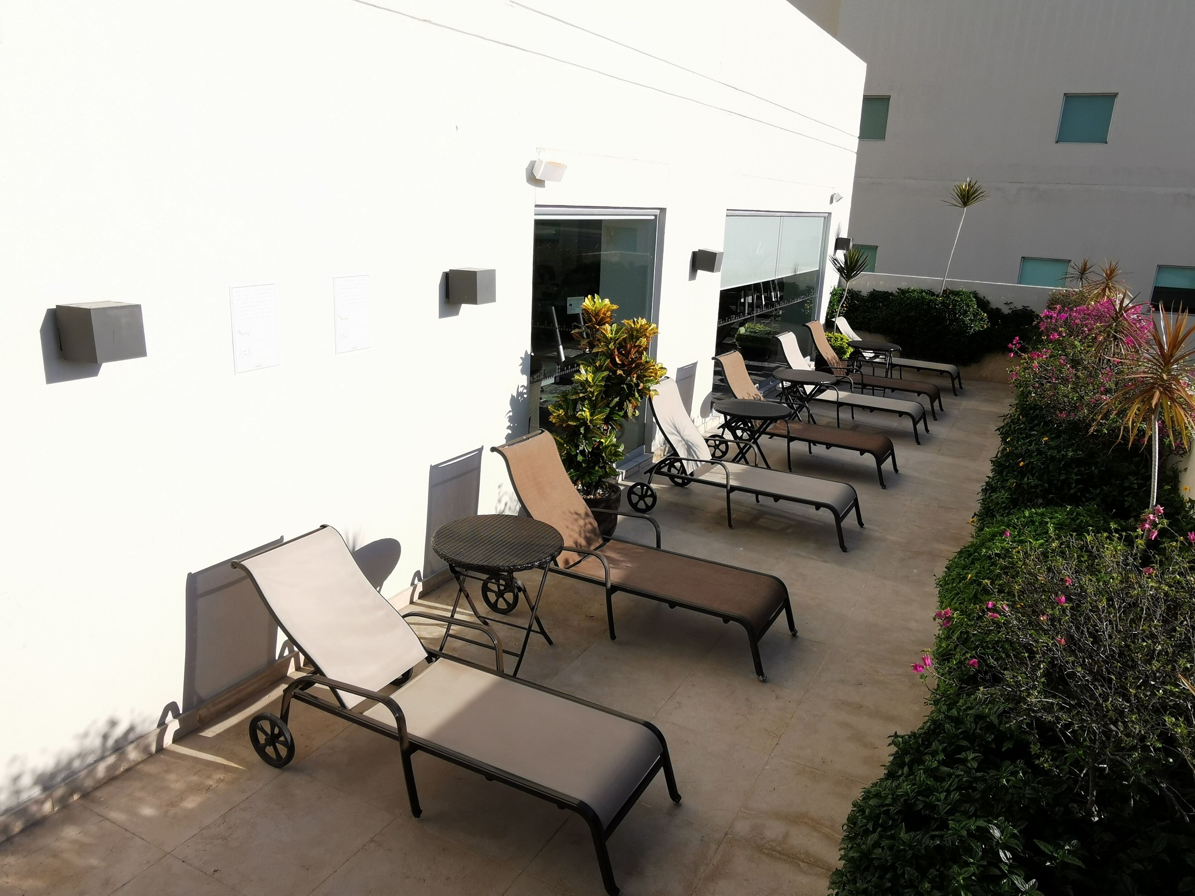 A sunlit outdoor seating area with lounge chairs and a small table, surrounded by greenery and a building facade.
