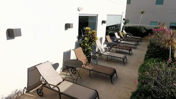 Un espace extérieur ensoleillé, aménagé avec des fauteuils confortables et une petite table, entouré de verdure et bordé par la façade d’un bâtiment.