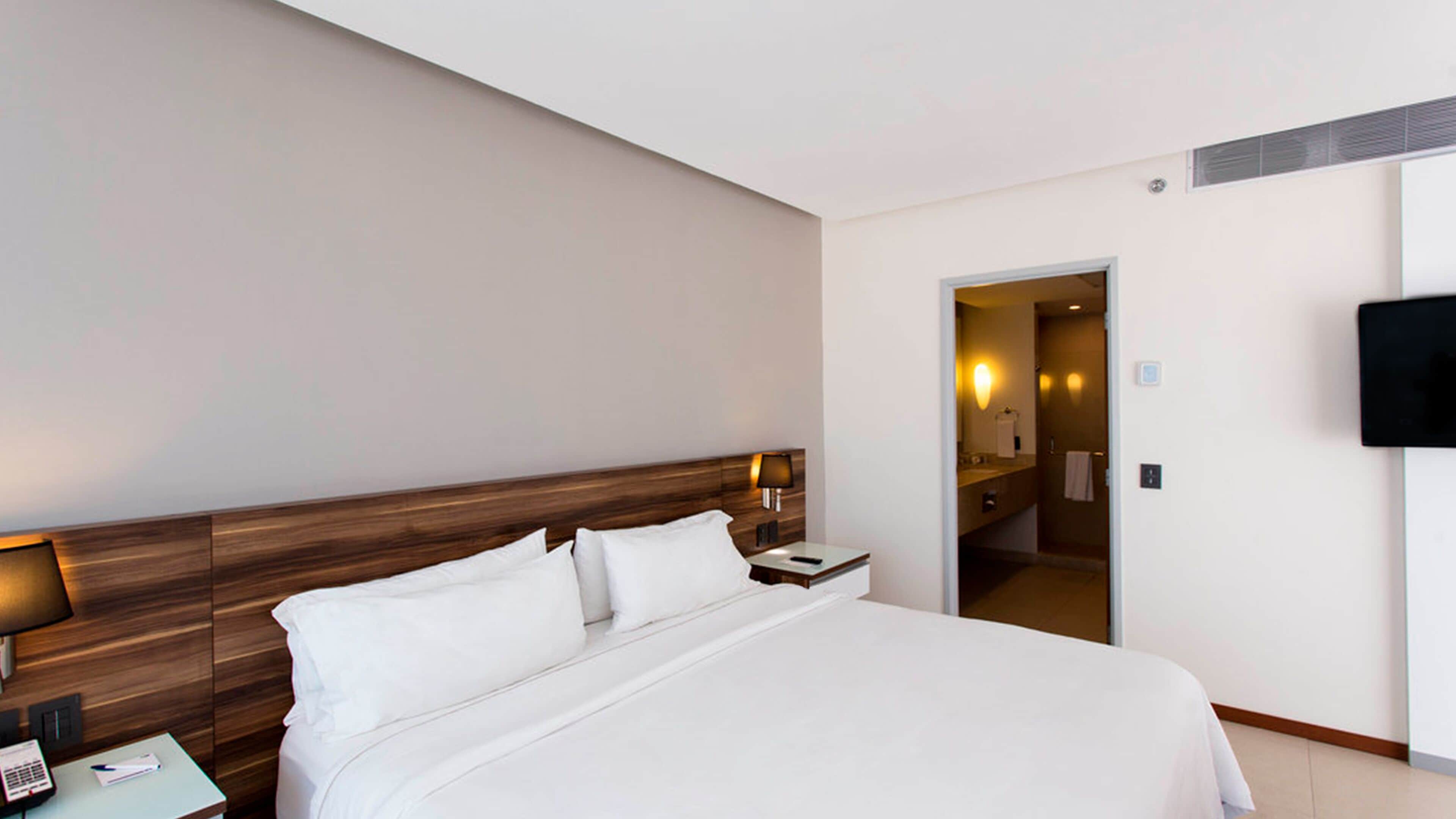 Een hotelkamer met een groot bed, een nachtkastje met een lamp, een telefoon en een aan de muur bevestigde televisie.
