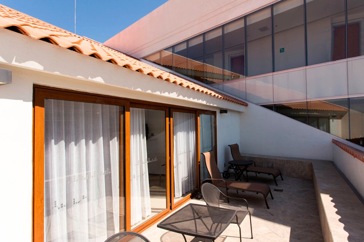 A sunlit outdoor patio with a table and chairs, a glass balustrade, and a view of a building with large windows.