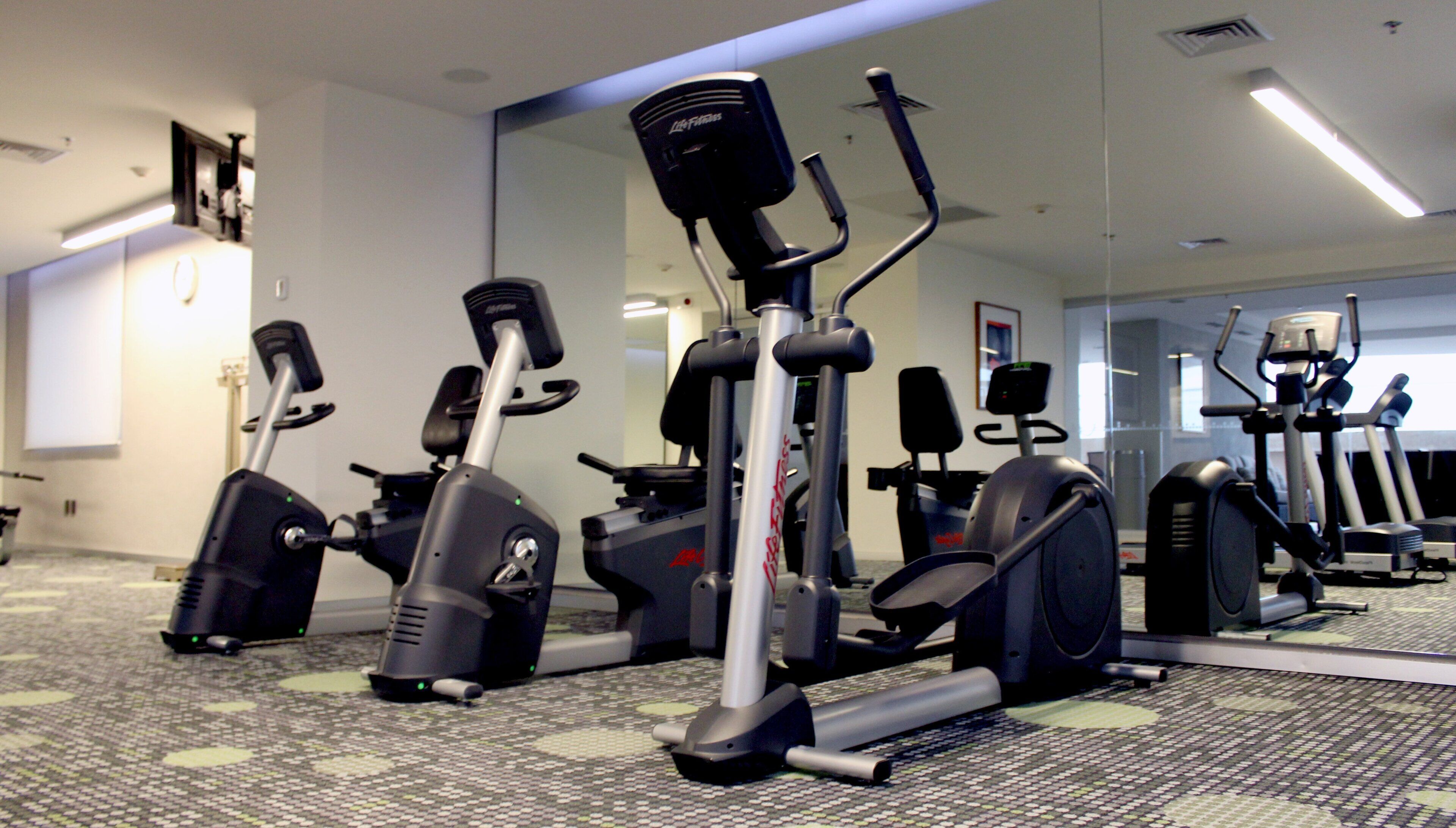 Ein Fitnessstudio mit verschiedenen Trainingsgeräten, einem Teppichboden und einem großen Spiegel, der die Geräte reflektiert.