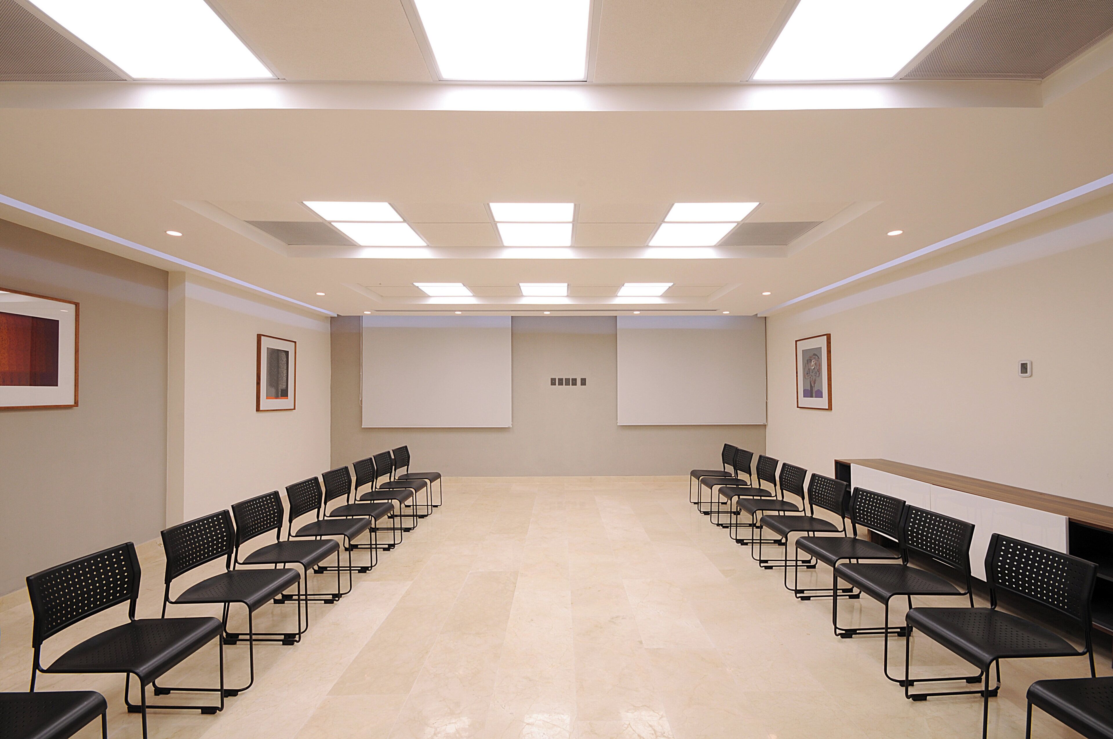 Sala de conferências com fileiras de cadeiras pretas, uma tela de projeção grande e luminárias no teto.