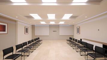 Sala de conferências com fileiras de cadeiras pretas, uma tela de projeção grande e luminárias no teto.