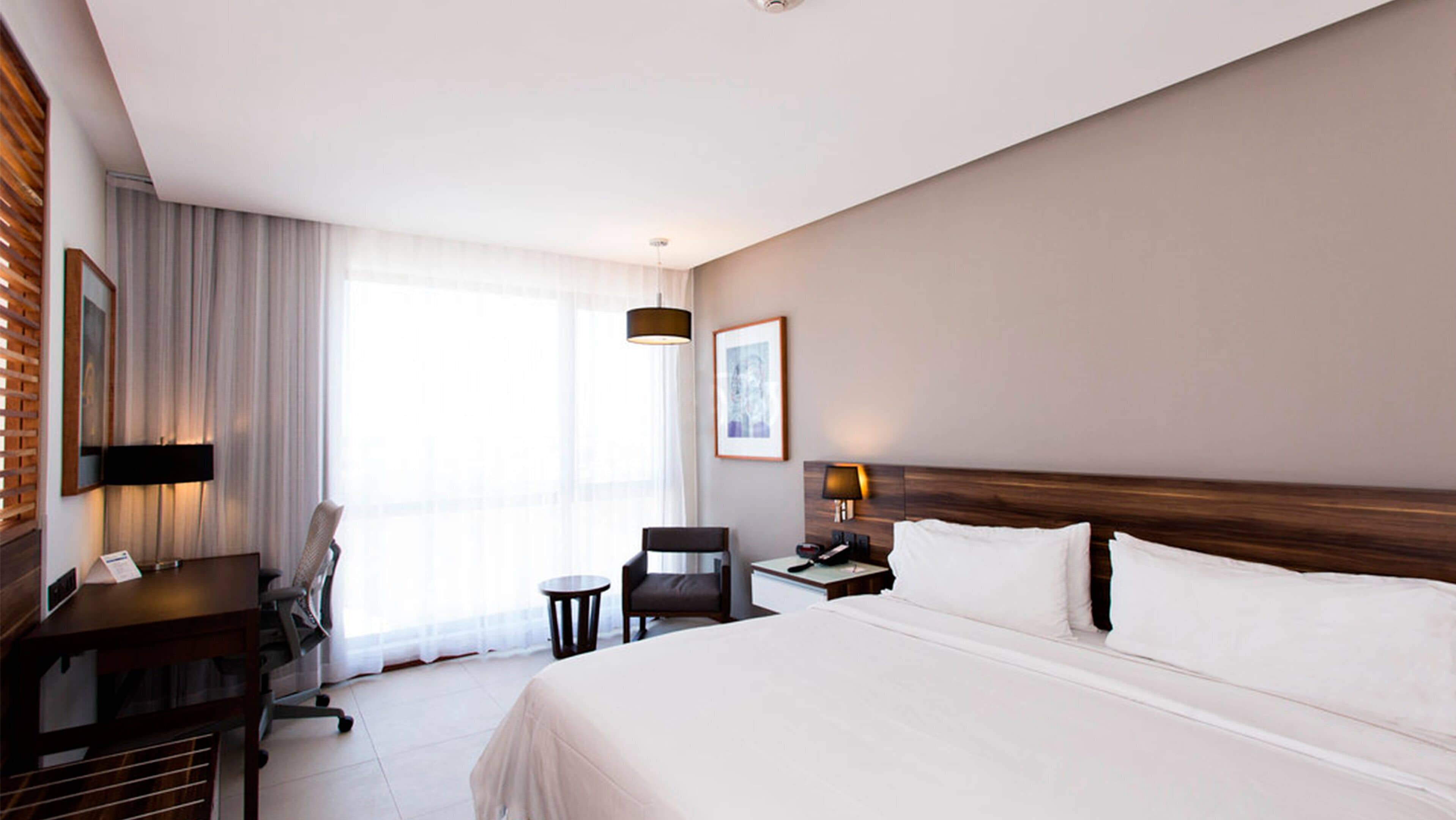 A hotel room with a large bed, a desk with a chair, a small table, a lamp, and a picture on the wall.