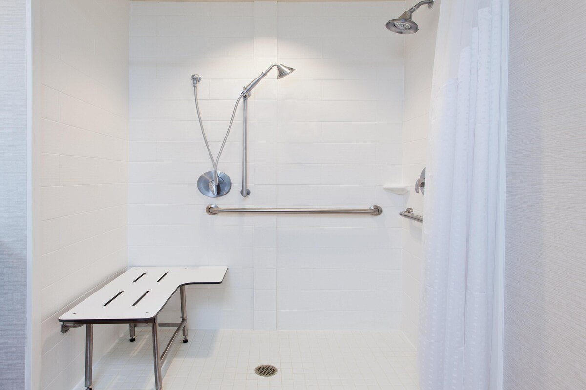 A modern bathroom with white tiles, a shower with a handheld showerhead, a grab bar, and a bench.