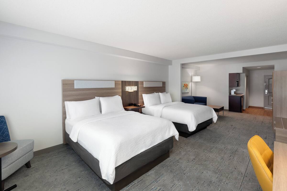Ein Hotelzimmer mit zwei Betten, einem Schreibtisch, einem Stuhl und einer kleinen Küchenzeile.