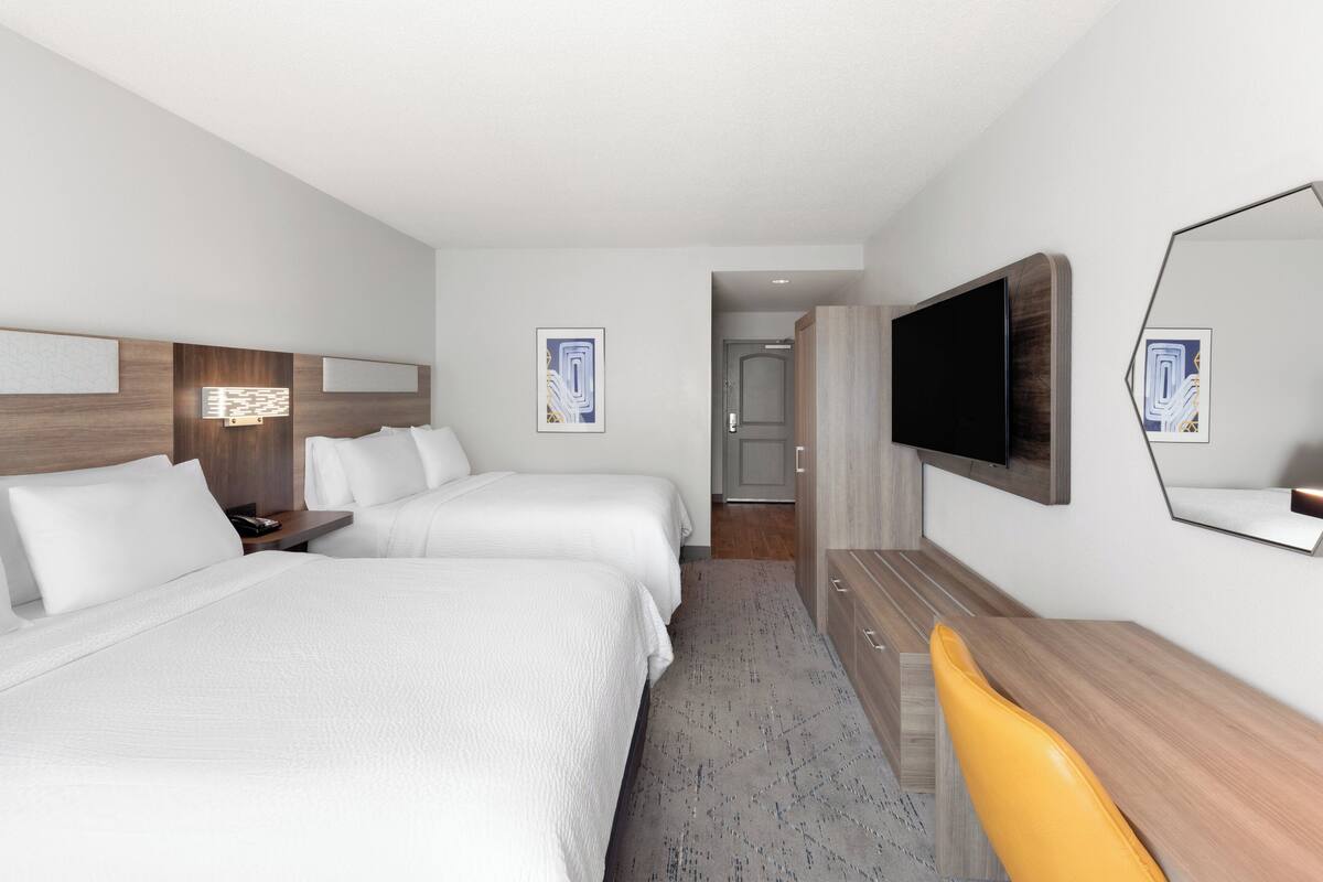 Ein Hotelzimmer mit zwei Betten, einem Schreibtisch, einem Sessel, einem Fernseher und Bildern an den Wänden.