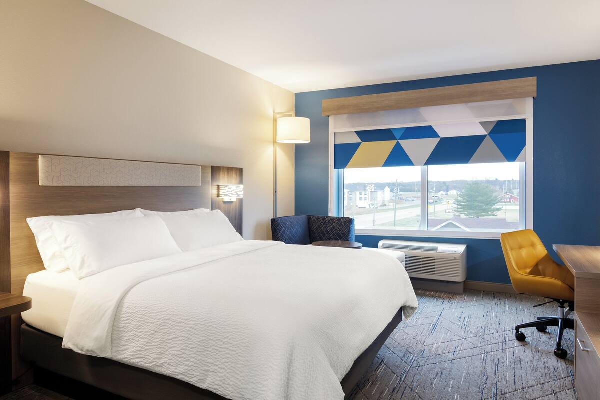 Ein Hotelzimmer mit einem großen Bett, einem Schreibtisch, einem Sessel und einem Fenster mit Blick auf die Stadt.