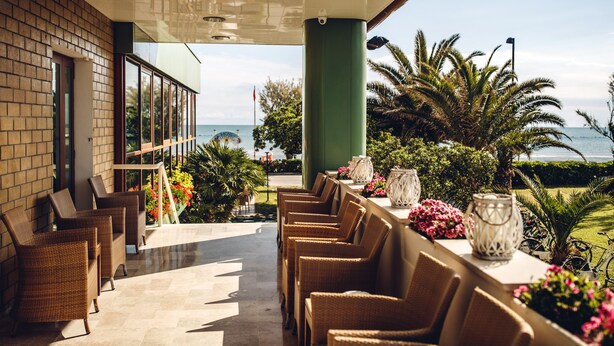 A terrace with wicker chairs and a table, overlooking a beach with palm trees and a view of the ocean. - UNAWAY Imperial Beach Fano (Fano)