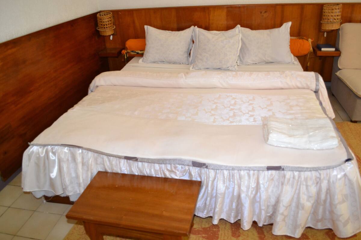 A large bed with a wooden headboard, two bedside tables with lamps, a bench, and a white bedspread.