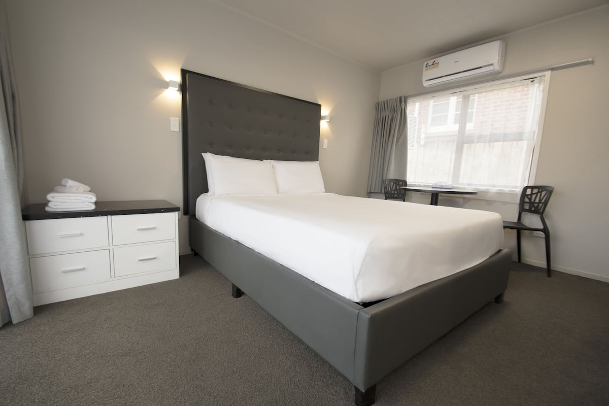 A neatly arranged hotel room with a bed, a bedside table, a chair, and an air conditioning unit.