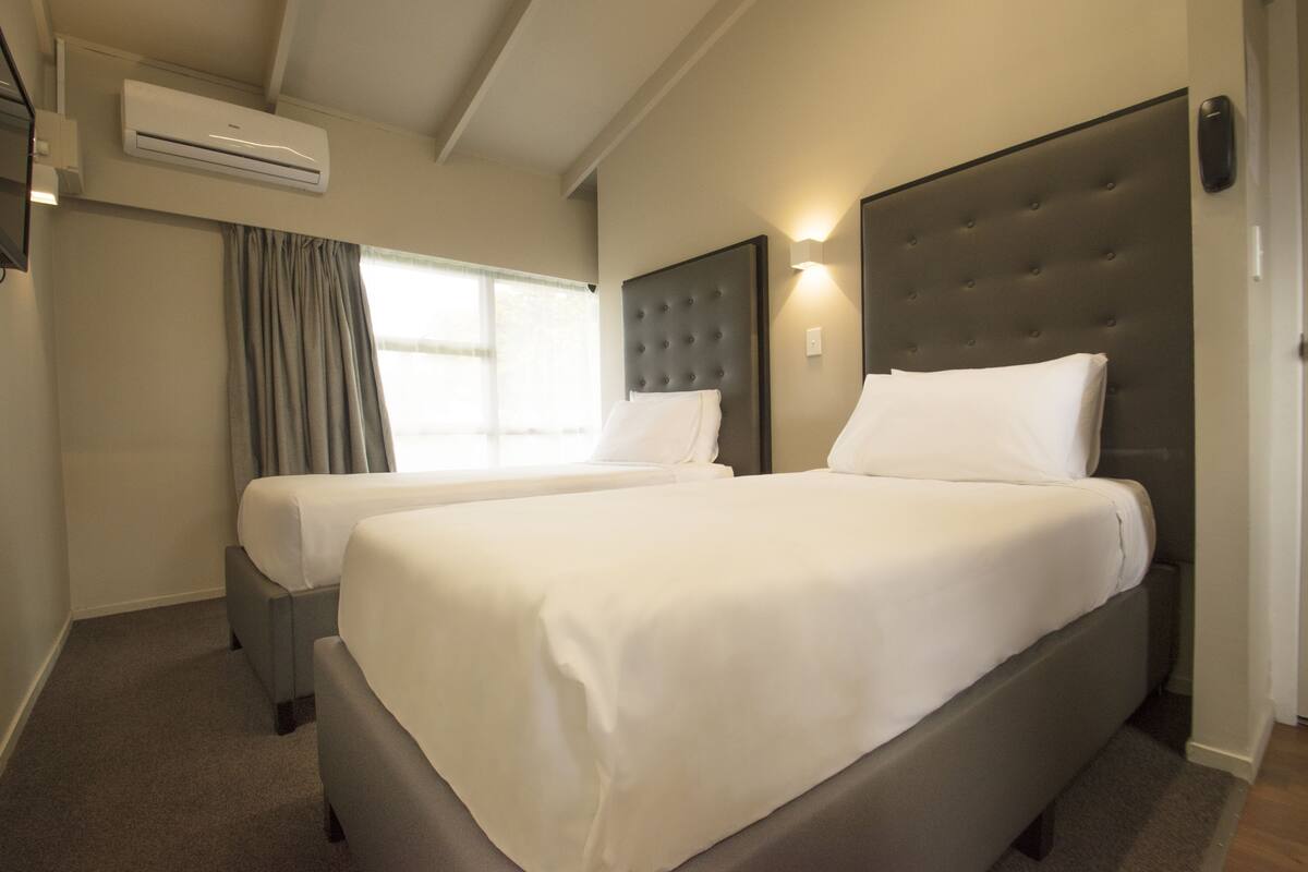 A hotel room with two beds, a large padded headboard, a window with curtains, and an air conditioning unit.