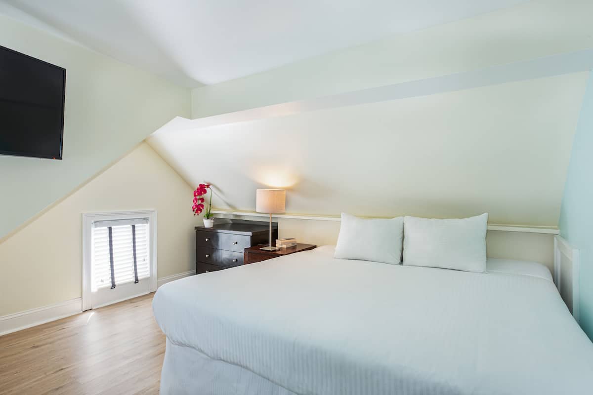A bedroom with a large bed, a nightstand with a lamp, a small window with blinds, and a wall-mounted TV.