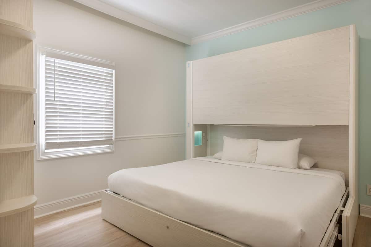 A bedroom with a bed, a window with blinds, a built-in shelving unit, and a light-colored wall.