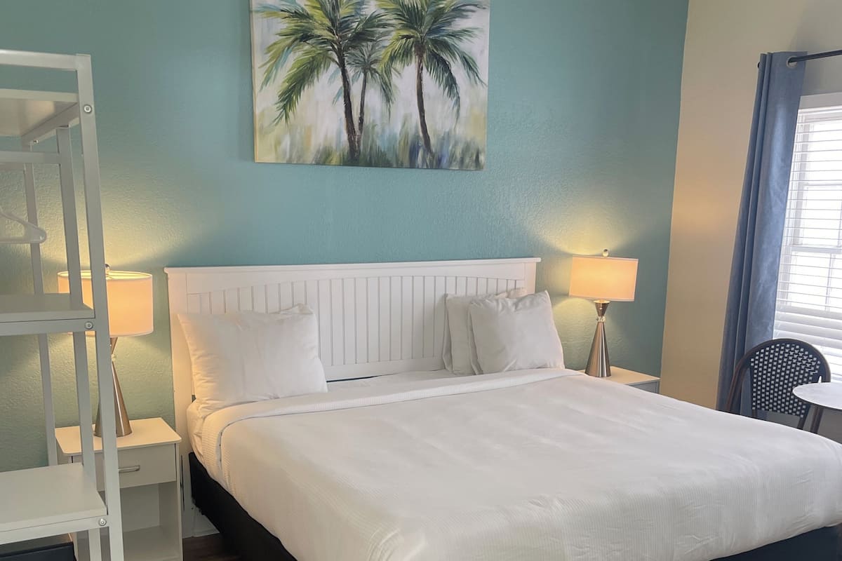 A bedroom with a bed, nightstands, a lamp, a painting of palm trees, and a wooden floor.