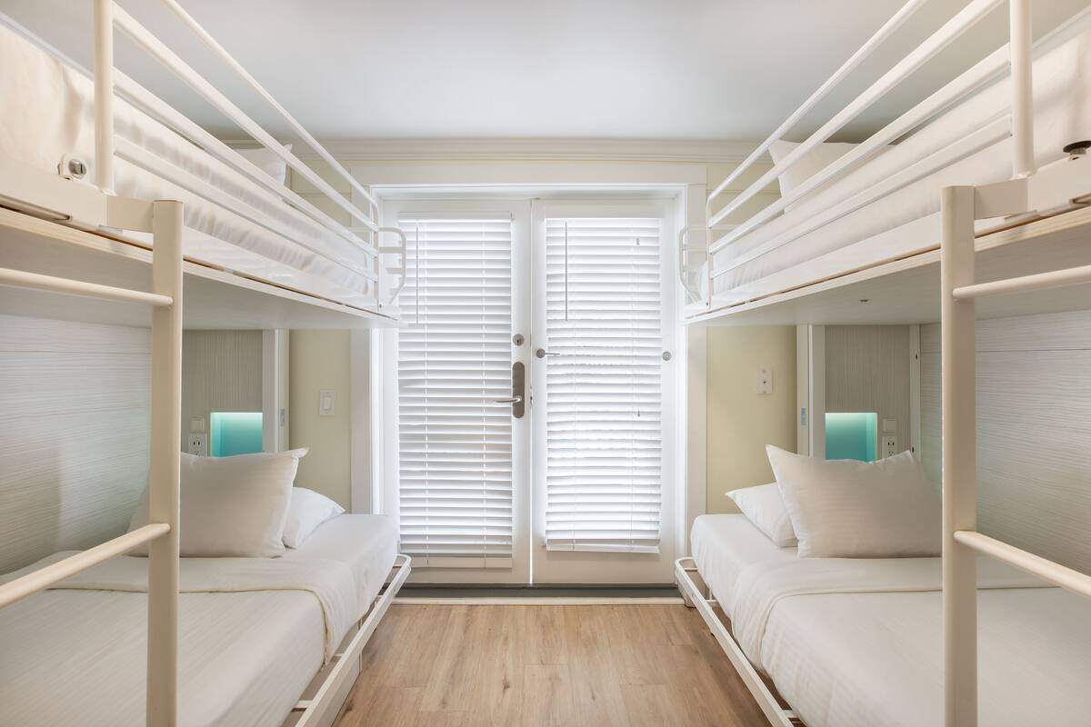 A room with bunk beds, a door with blinds, and a bedside table with a lamp.