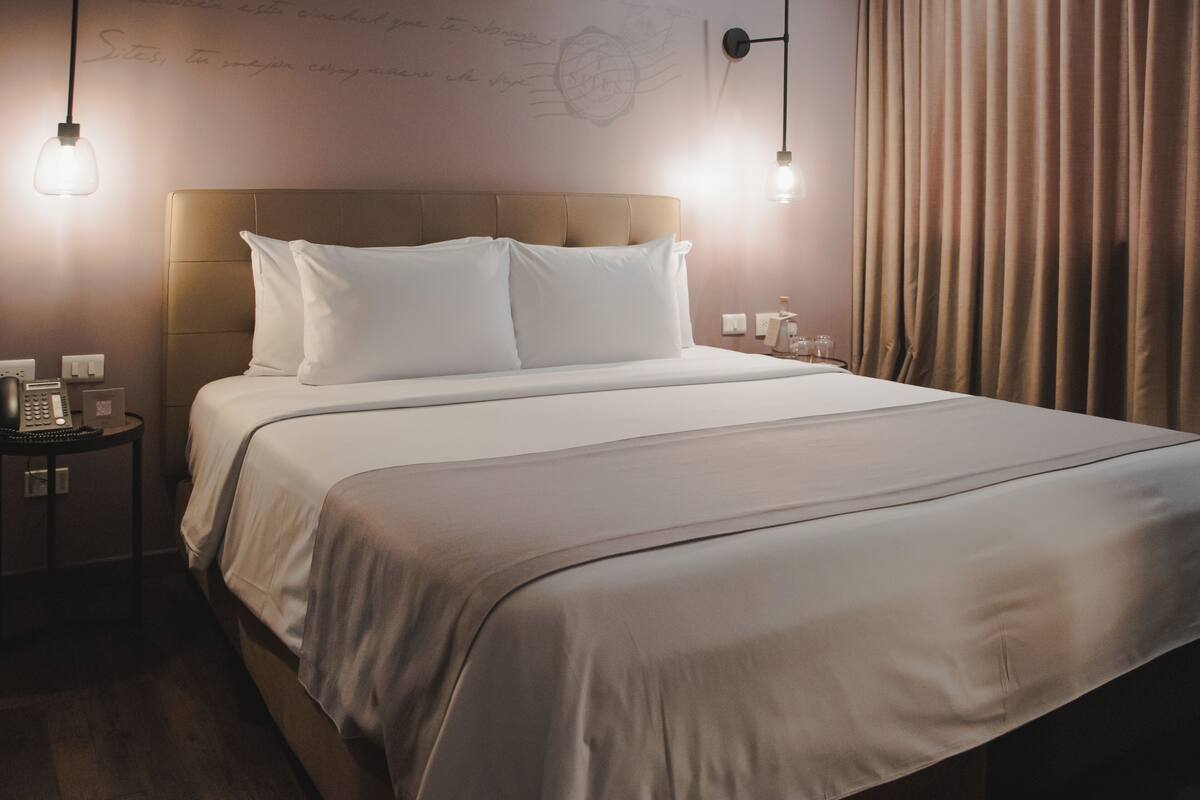 A neatly made bed with white linens and a headboard, a bedside table with a lamp, and a wall with a decorative element.