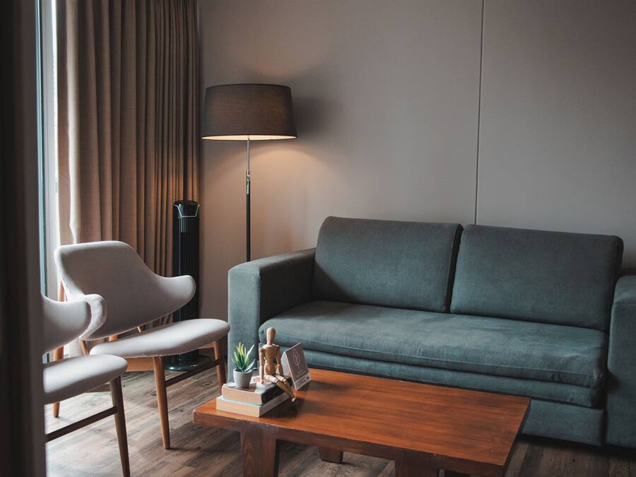 Uma sala de estar moderna com um sofá cinza, uma mesa de centro de madeira e uma poltrona branca. Uma luminária de chão e um vaso com planta também estão visíveis.