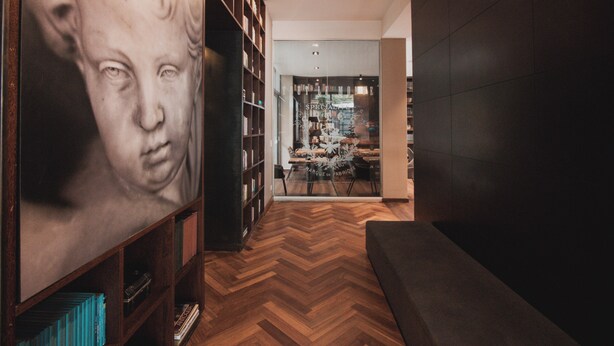 A hallway with a wooden herringbone floor, a large framed artwork on the left, and a seating area on the right. - Sites Hotel (Medellín)