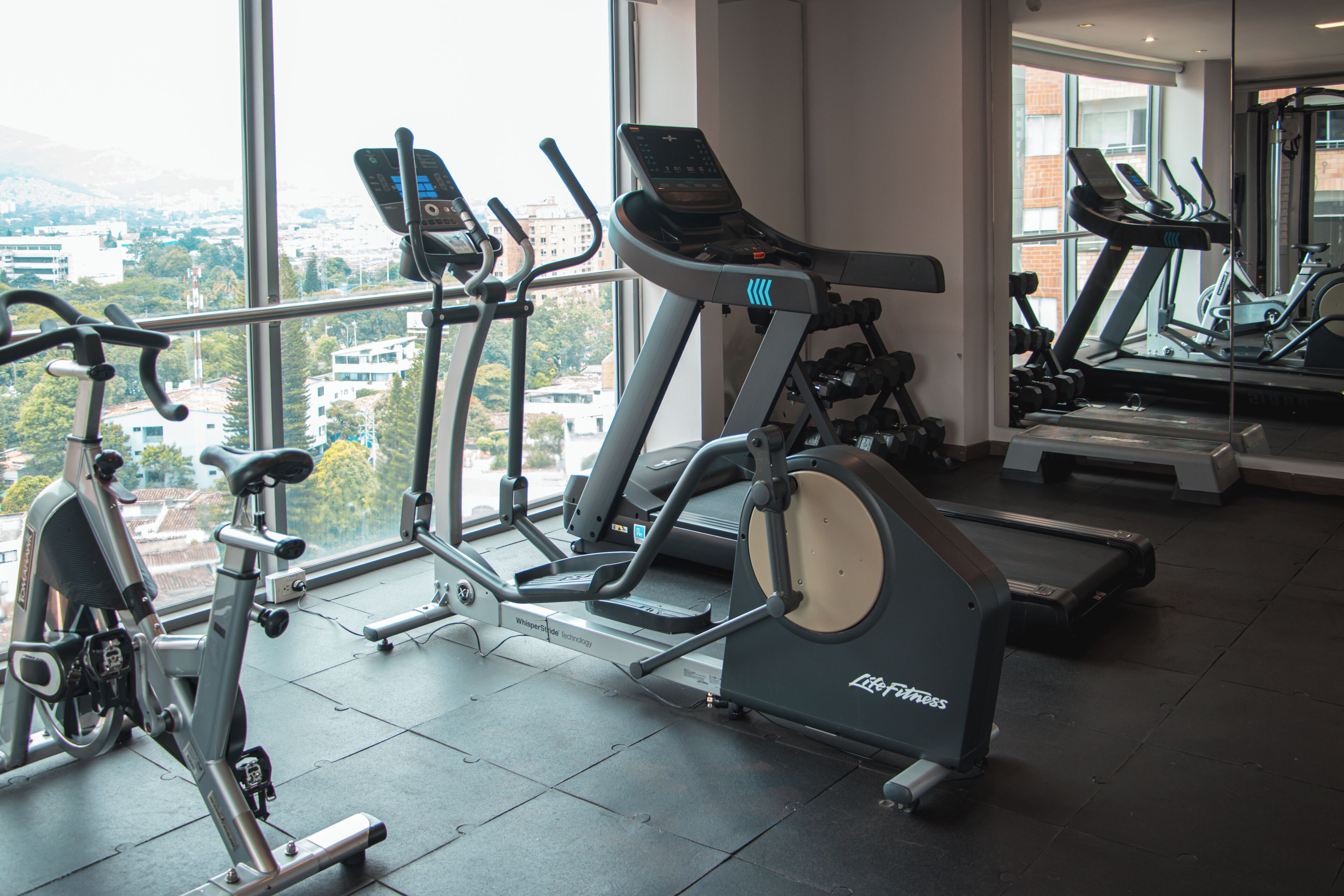 Un gimnasio con equipos de ejercicio variados, amplias ventanas y vista a la ciudad.