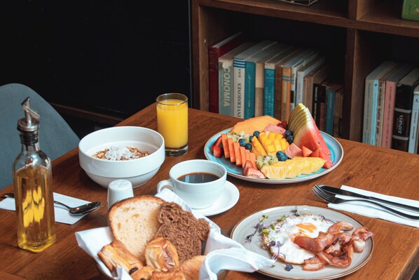 A breakfast table with a bowl of cereal, a glass of orange juice, a plate of fruit, toast, and a plate of bacon and eggs. - Sites Hotel (Medellín)