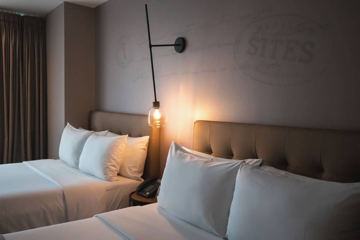 A hotel room with two beds, a nightstand, a lamp, and a wall with the word "SITES" partially visible.
