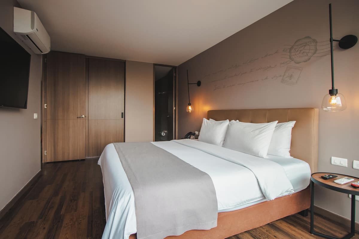 A modern hotel room with a large bed, wooden flooring, a flat-screen TV, and a wall-mounted air conditioner.