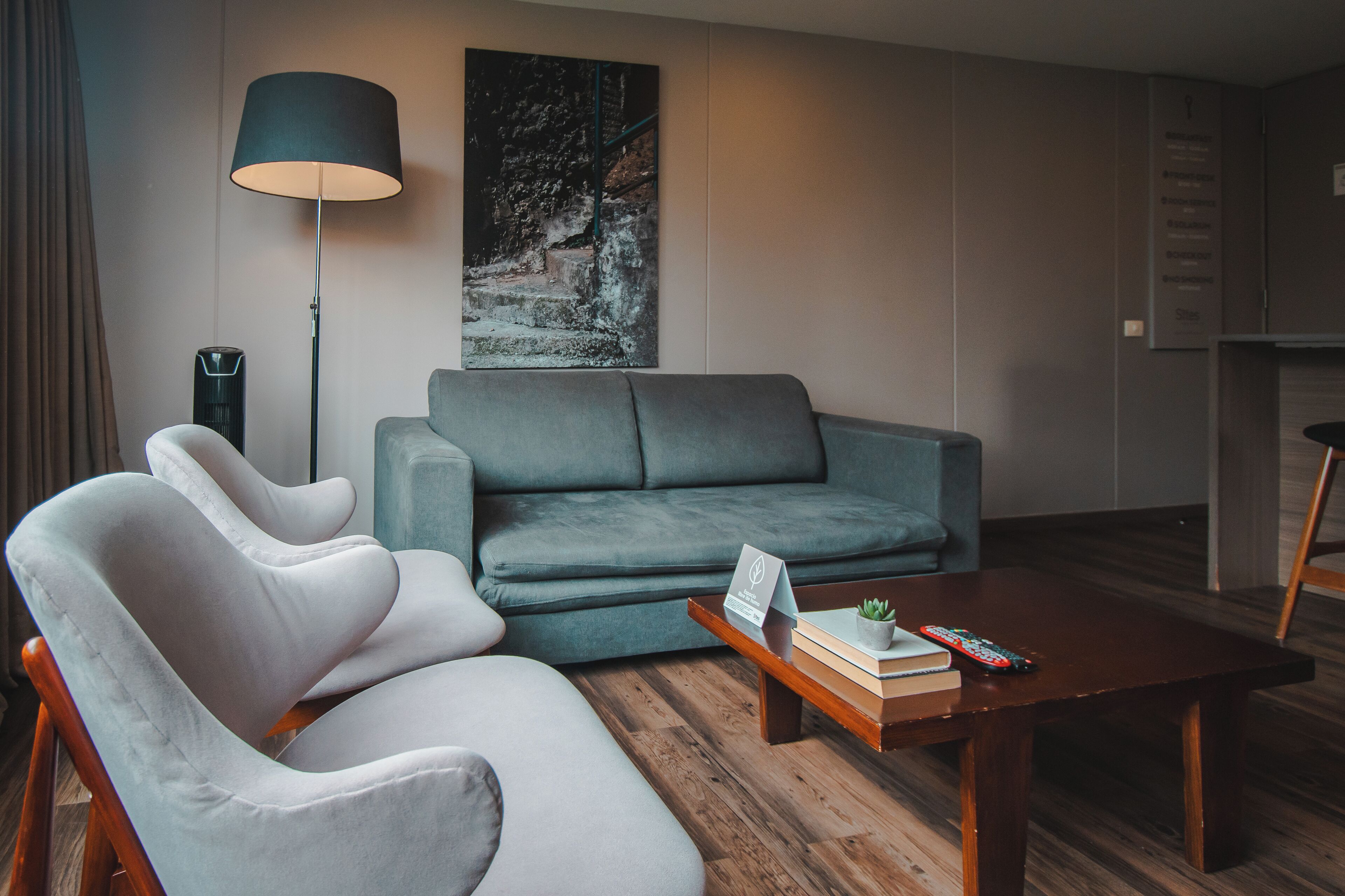Un salon moderne avec un canapé gris, un fauteuil blanc, une table basse en bois sur laquelle sont posés des livres et une télécommande, un lampadaire et un tableau accroché au mur.