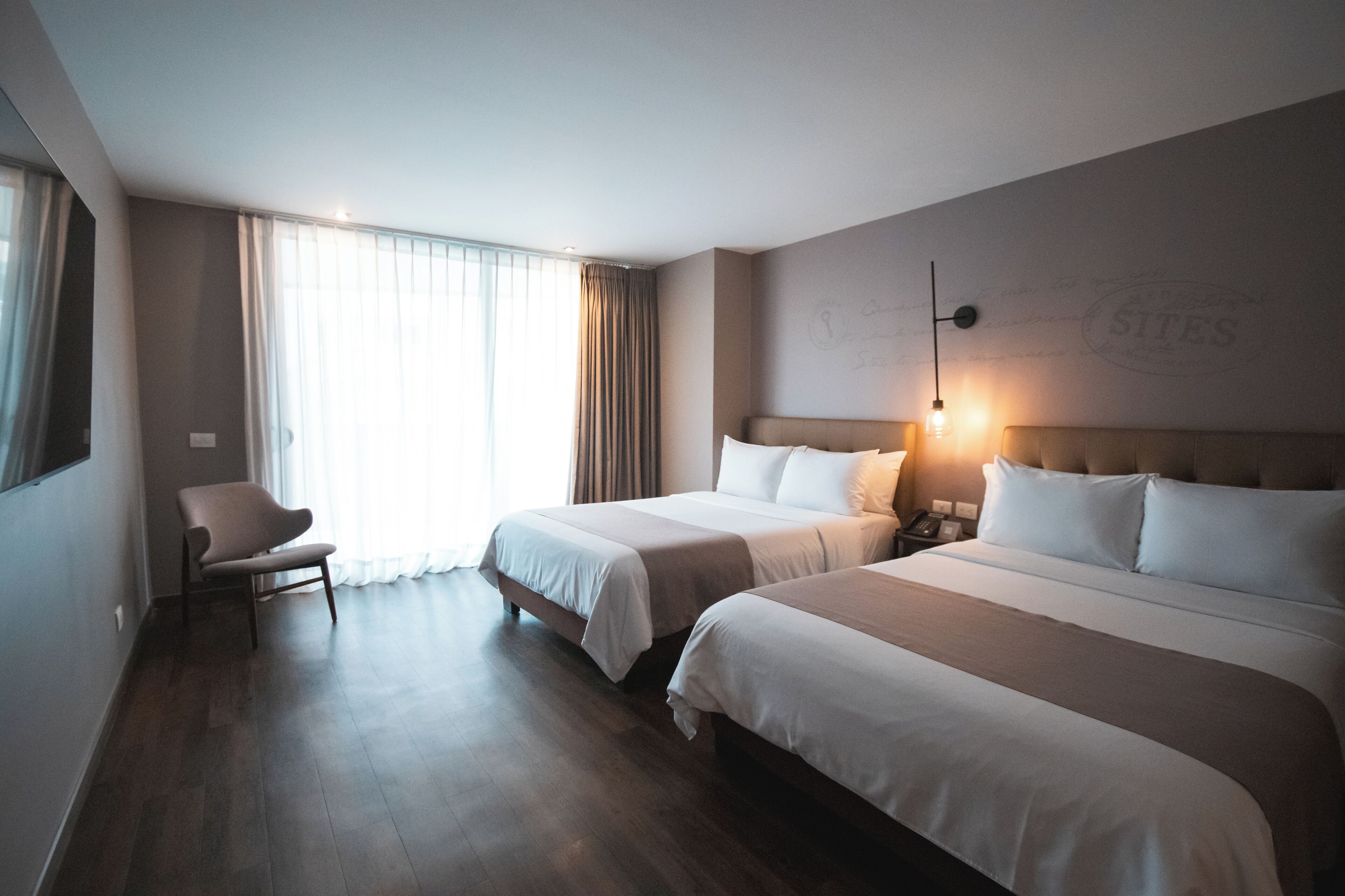 A hotel room with two beds, a chair, a bedside table with a lamp, and a large window with curtains.
