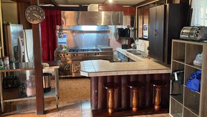 A kitchen with a stainless steel stove, a microwave, and a toaster. There is a clock on the wall and a red curtain. - Hostel La Choza Inn (La Fortuna)