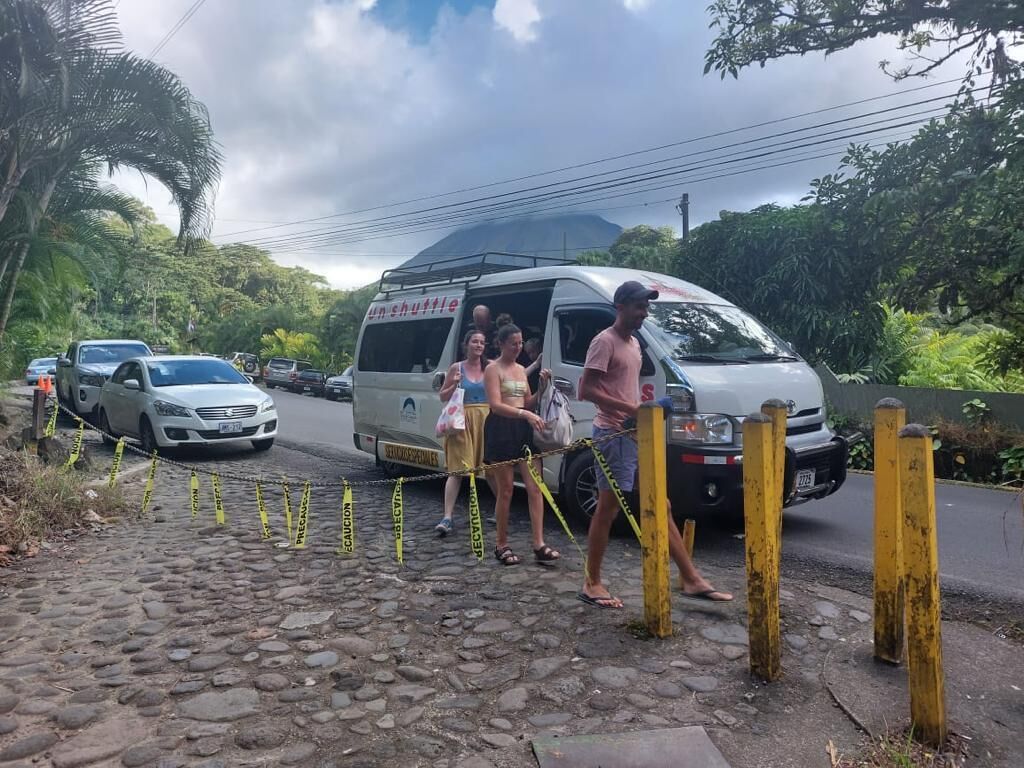 A shuttle bus parked on a cobblestone road with yellow tape, surrounded by greenery and a cloudy sky.