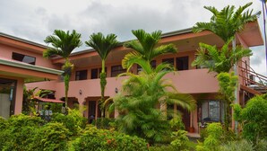 A multi-story building with a pink facade, surrounded by lush greenery and palm trees. - Hostel La Choza Inn (La Fortuna)