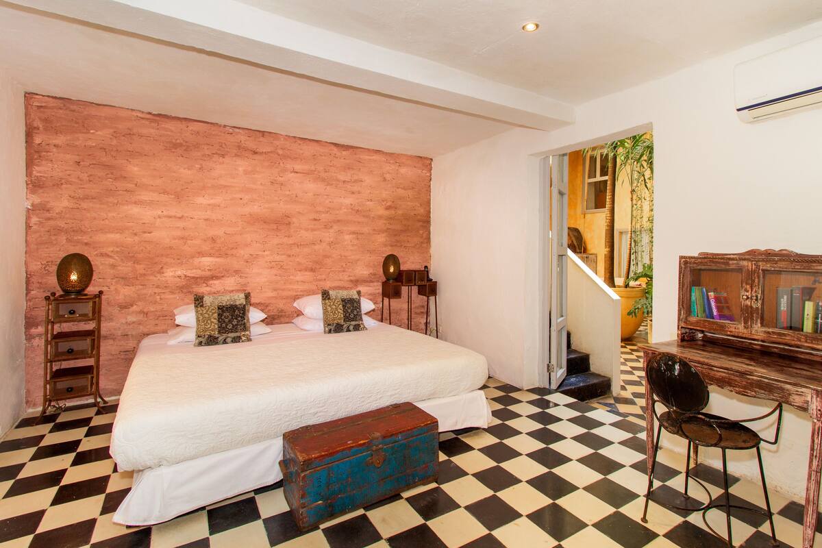 Slaapkamer met een geruite vloer, een groot bed, een houten kist, een bureau met boeken en een spiegel.