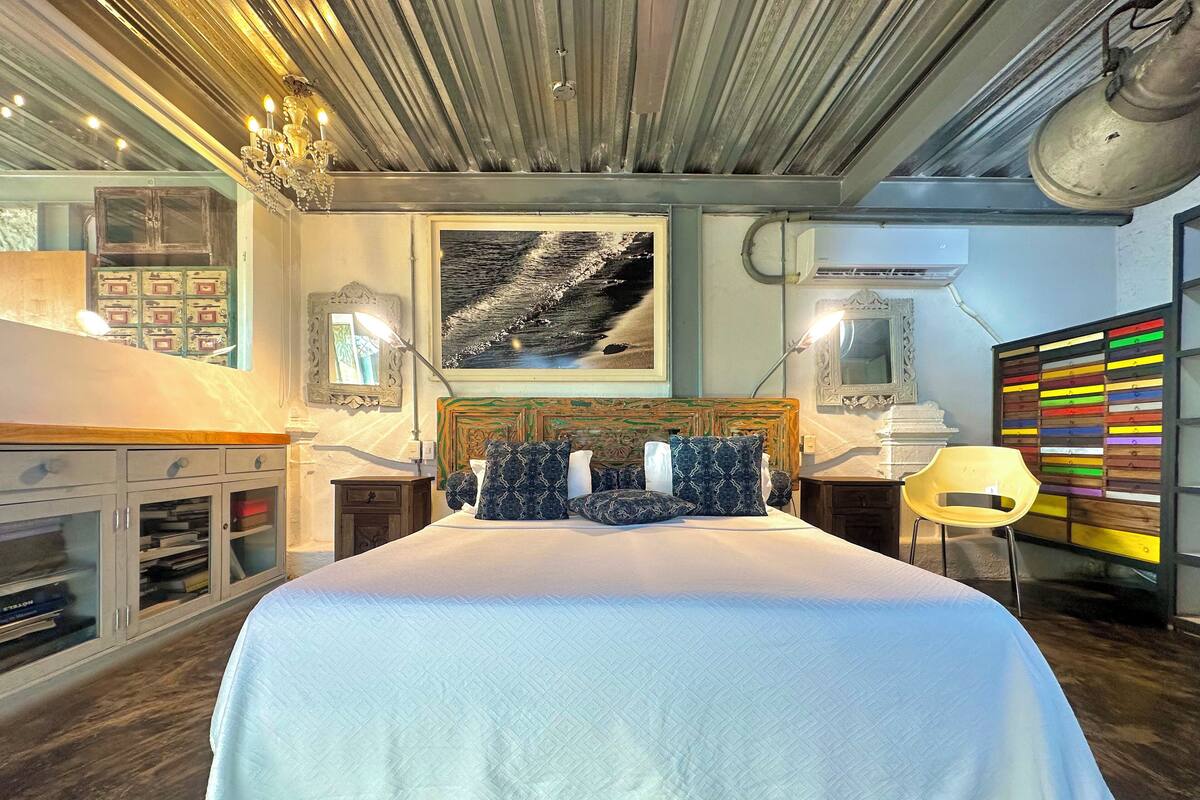 Een slaapkamer met een groot bed, een houten hoofdbord, een kroonluchter, een schilderij aan de muur en een kleurrijke wandversiering.