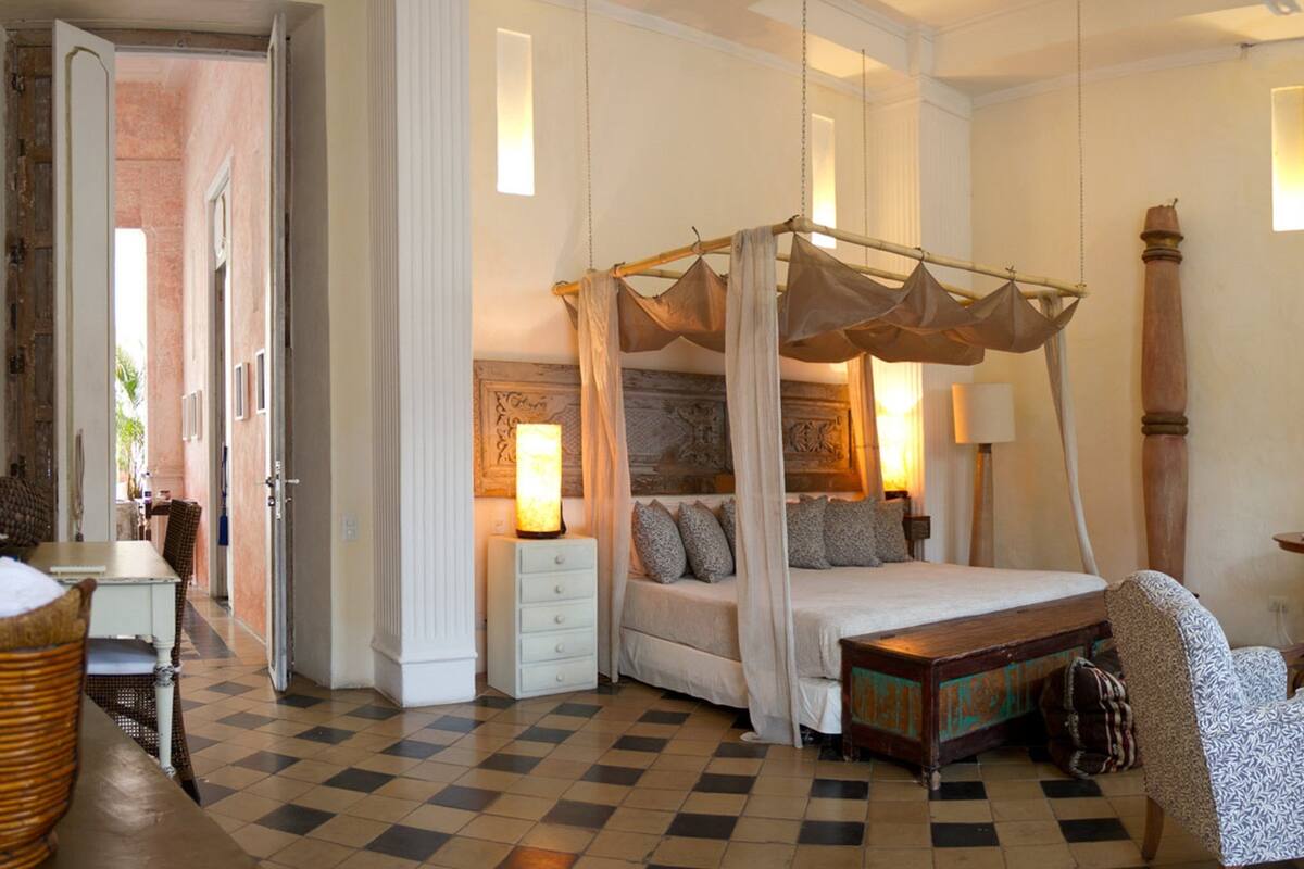 Ruime slaapkamer met een hemelbed, een zithoek met stoelen, een eettafel en een groot raam met gordijnen.