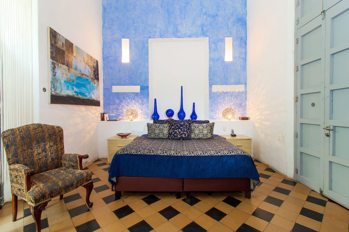 Een slaapkamer met een geruite vloer, een bed met een blauwe spreidek, een stoel en een muur met een schilderij en twee wandlampen.