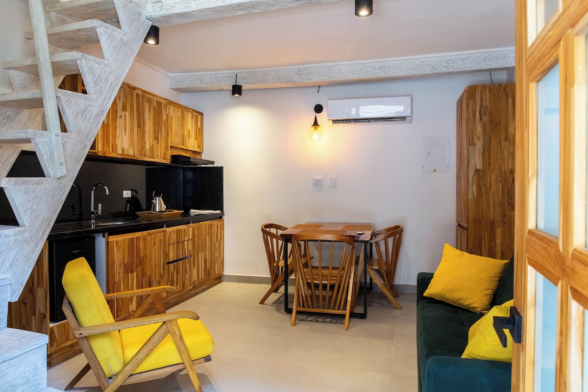 Een kleine keuken met houten kastjes, een eettafel met stoelen en een trap.