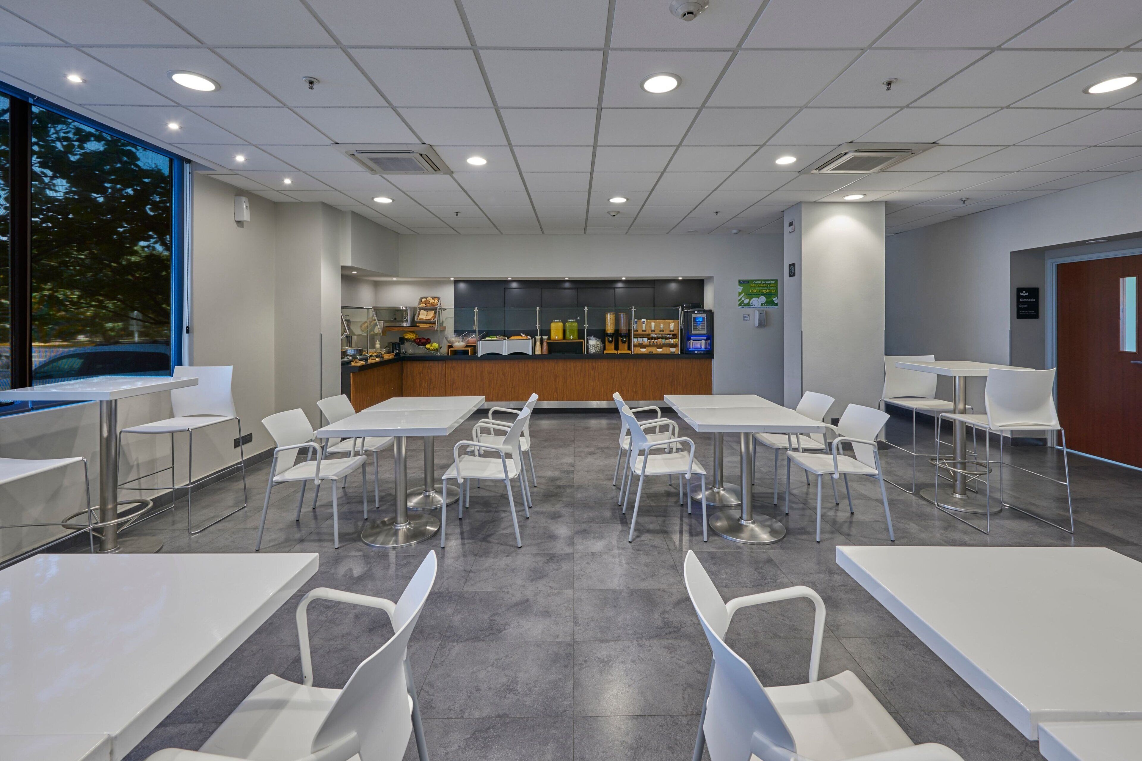 Una sala de conferencias moderna y bien iluminada, con mesas y sillas blancas, un área de recepción con mostrador y varios objetos, y ventanas grandes que ofrecen luz natural.