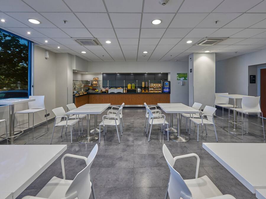 A modern, well-lit conference room with white tables and chairs, a reception area with a counter and various items, and large windows providing natural light.