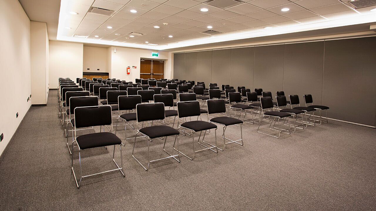 Un salón de conferencias con filas de sillas negras, un escenario y un extintor en la pared.