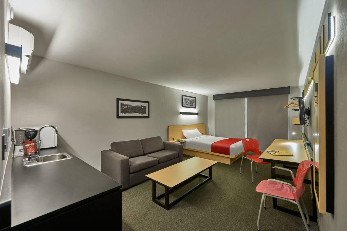 A compact hotel room with a kitchenette, a single bed, a sofa, a dining table, and a small desk.