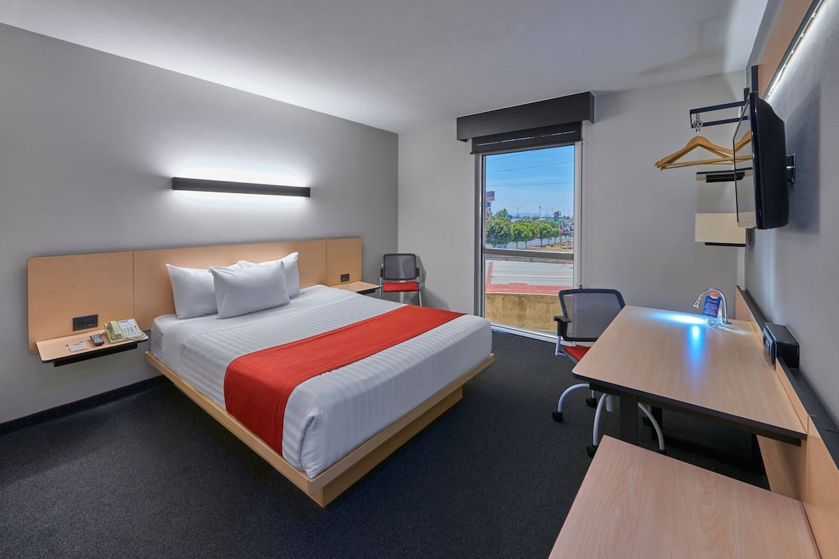 A hotel room with a bed, a desk, a chair, a TV mounted on the wall, and a window with a view of the outside.