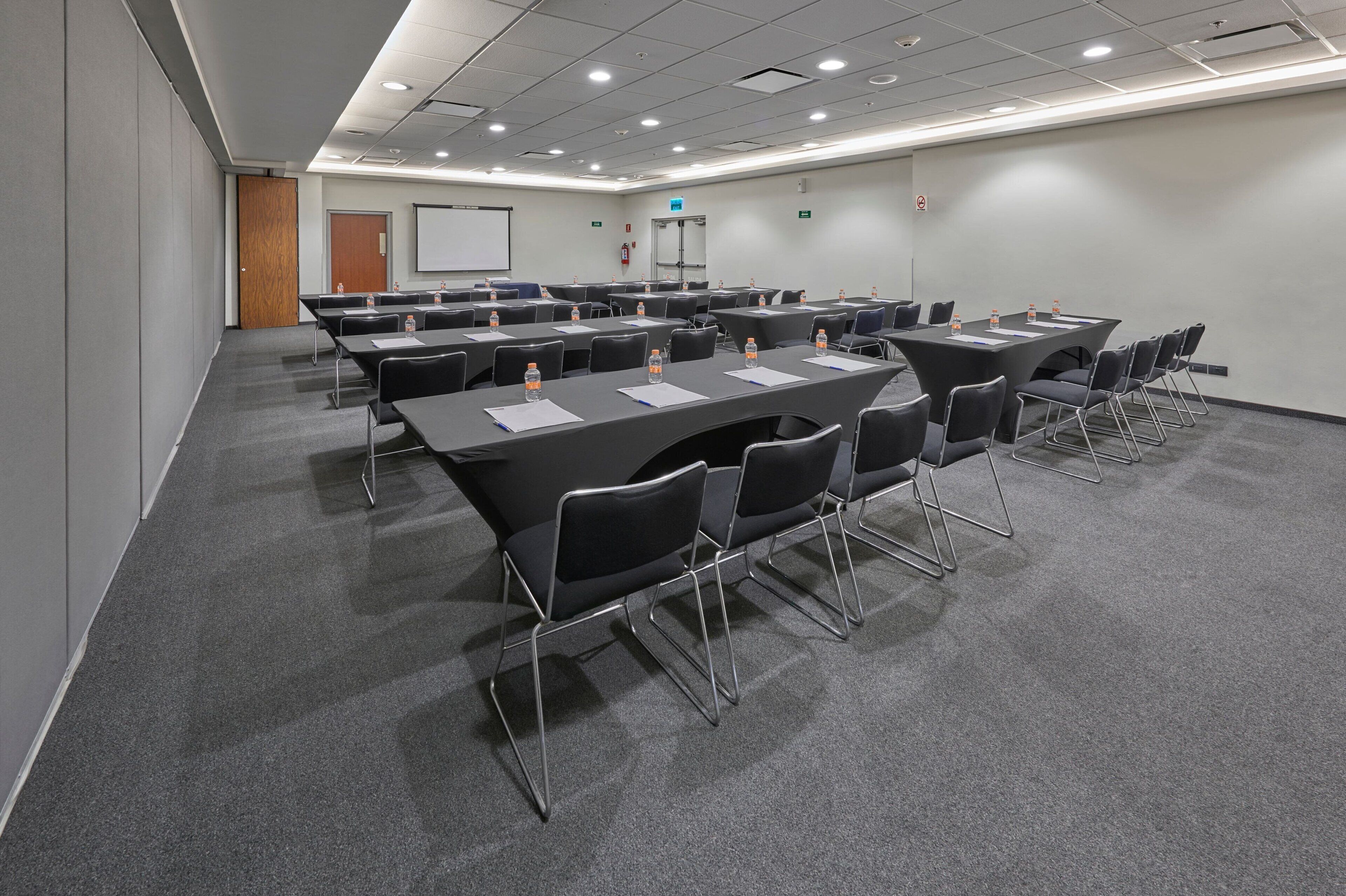 Un salón de conferencias con varias mesas y sillas, equipado con botellas de agua y documentos, y una pizarra en la pared.