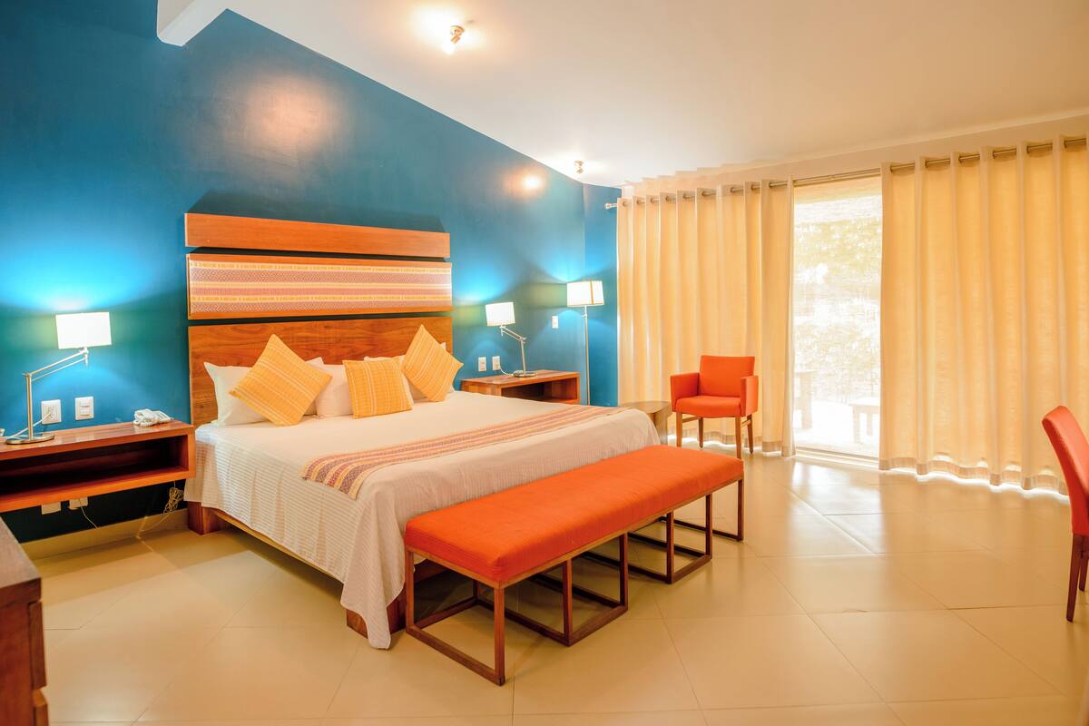 A modern bedroom with a large bed, a bench, two nightstands, a chair, and a view of the outdoors.