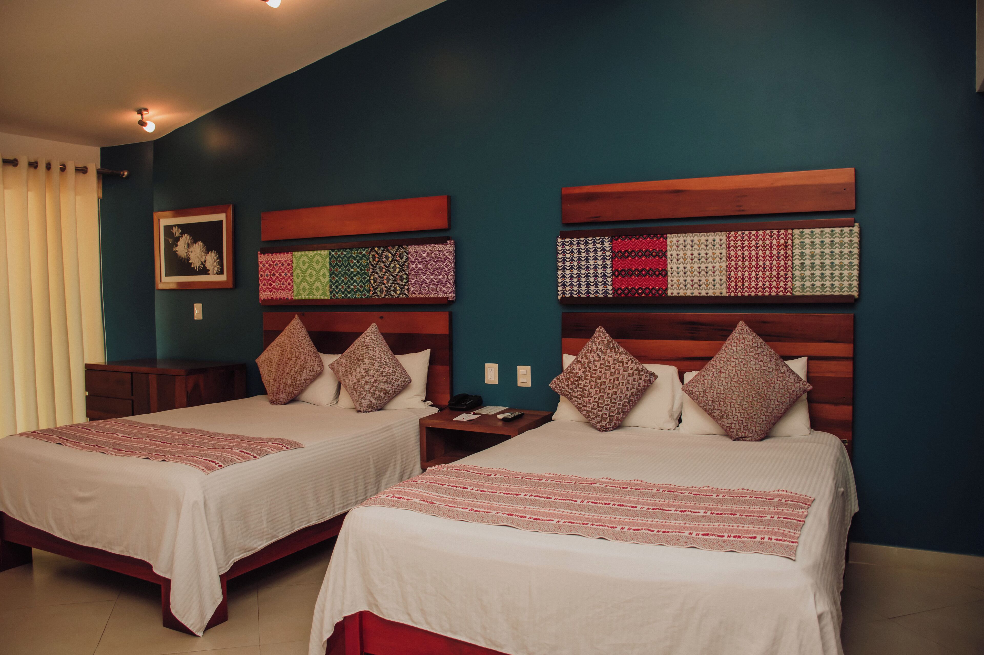Habitación de hotel con dos camas, cada una con cabecero de madera, ropa de cama blanca y una manta con rayas rojas. Entre las camas hay una mesita de noche con un teléfono y una lámpara.